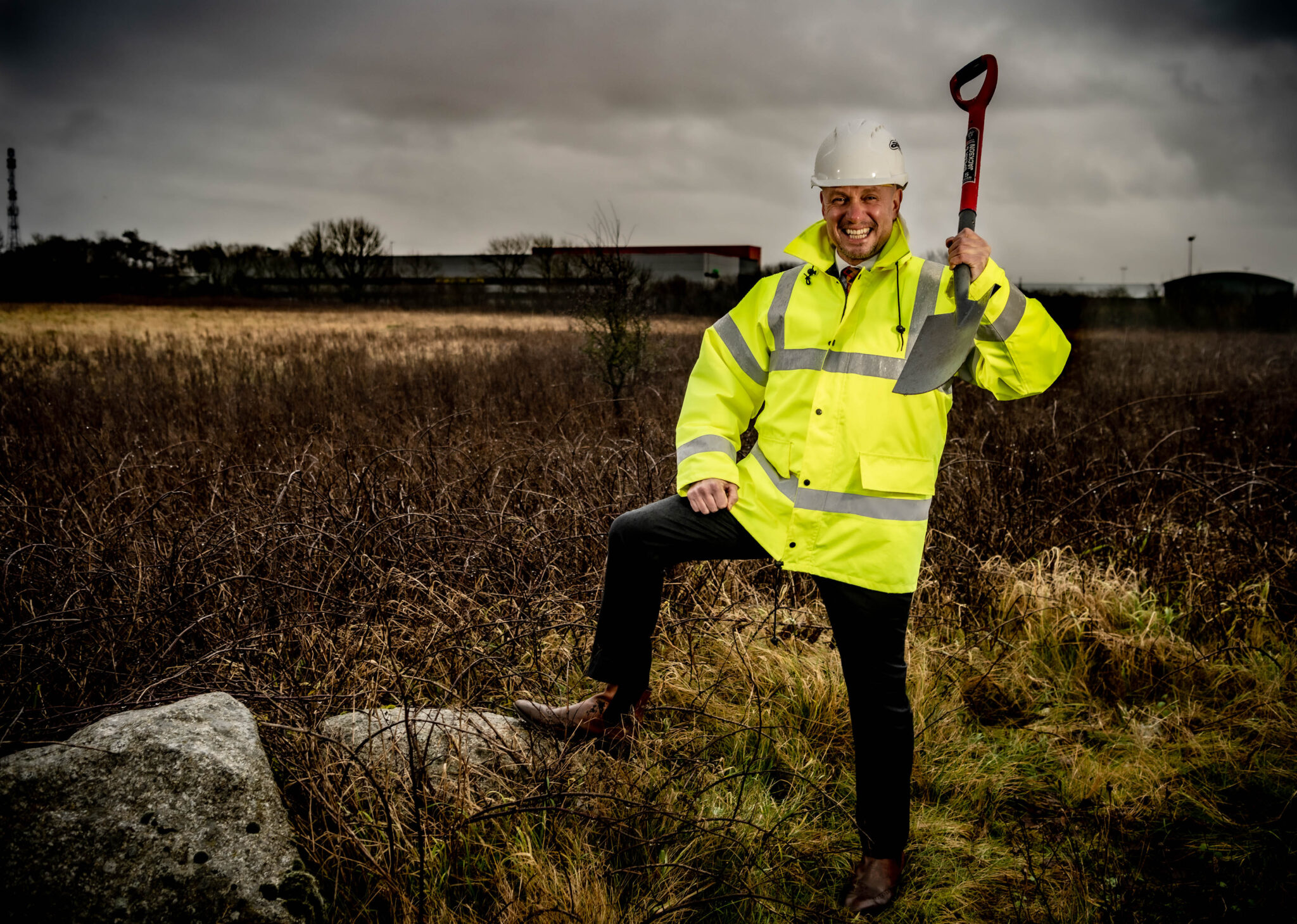 Chris Nattress, Lakes College principal, sports a hi-vis jacket and hard hat as he celebrates the approval of the college's new multi-million pound Civil Engineering Training Centre. He is seen at the site of the new build, holding a spade and grinning.