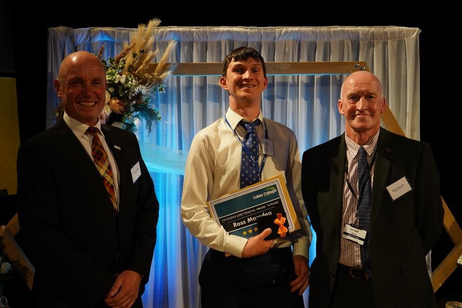 A student holds a certificate with two men either side presenting him with an award